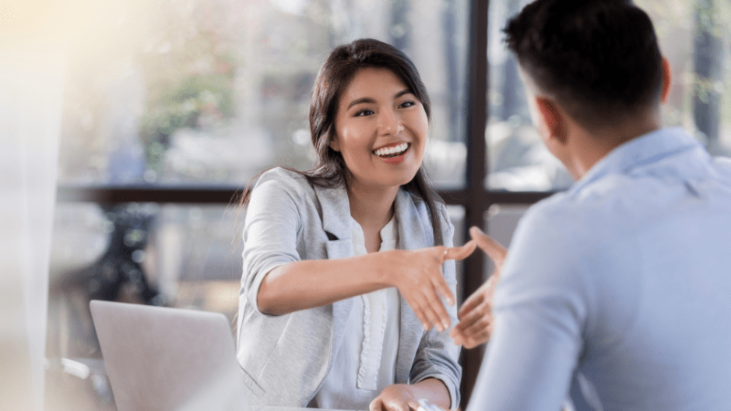 A smiling woman offers a handshake to a man seated opposite her at a job interview